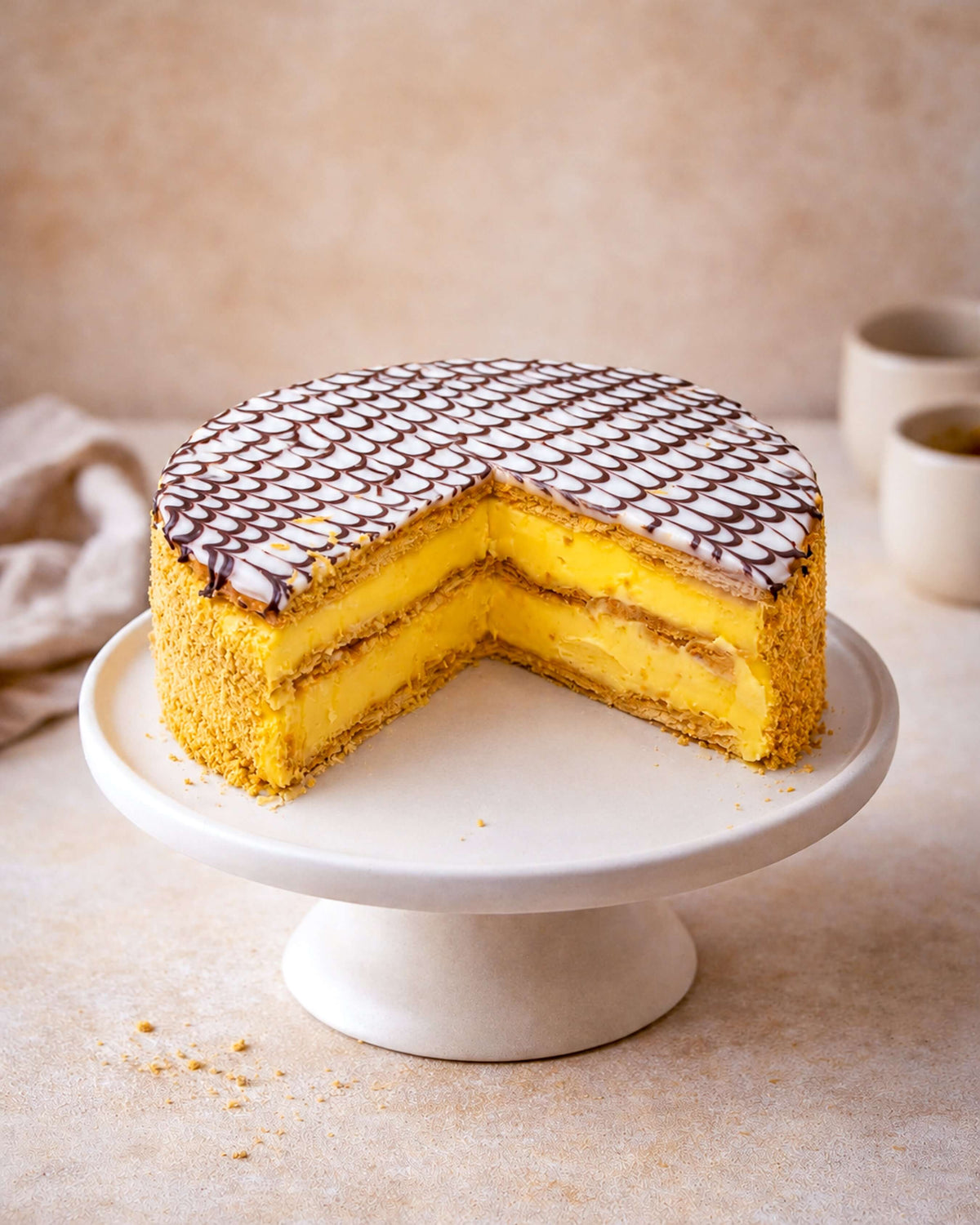 Mille Feuille Gateau with golden layers of cream and custard, topped with fondant and chocolate drizzle on a cake stand.